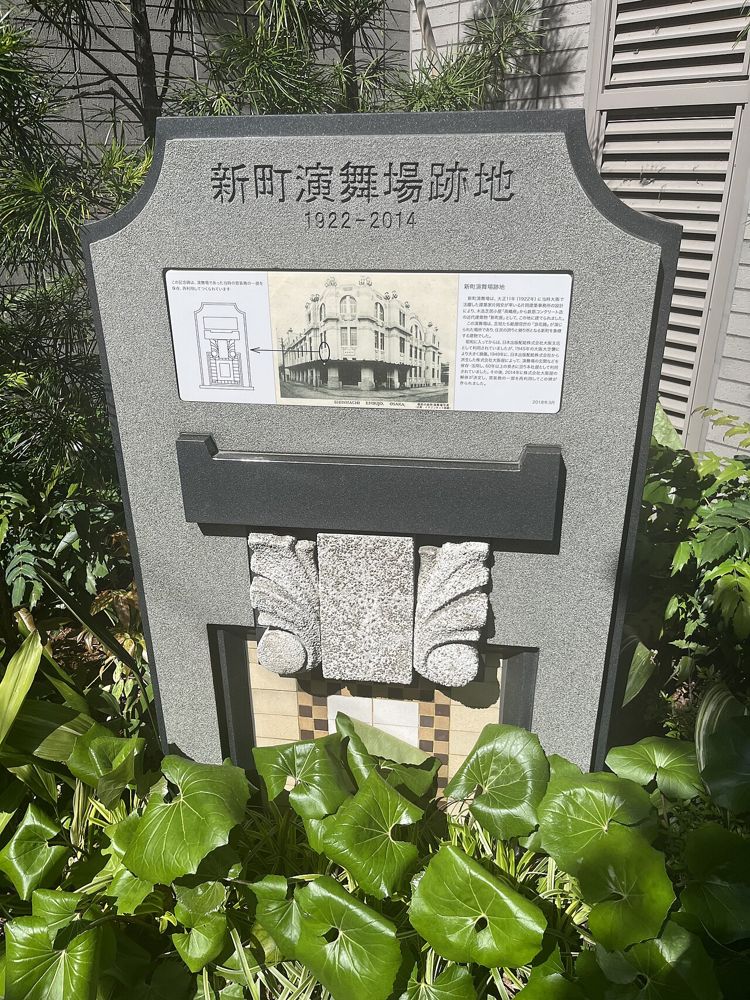 高層建物の脇の植え込みに立つ「新町演舞場跡地」の石碑と説明板